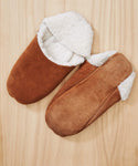 A pair of Suede Moroccan Slippers with white fluffy shearling linings rests on a light wooden surface. One slipper is shown facing up, while the other displays its sole.