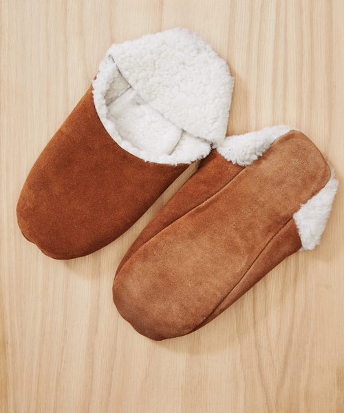 A pair of Suede Moroccan Slippers with white fluffy shearling linings rests on a light wooden surface. One slipper is shown facing up, while the other displays its sole.