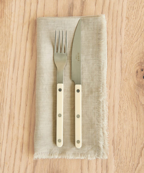 A fork and knife from the Bistro Vintage Finish Cutlery Set, featuring cream handles, rest side by side on a beige linen napkin with frayed edges atop a light wood table.