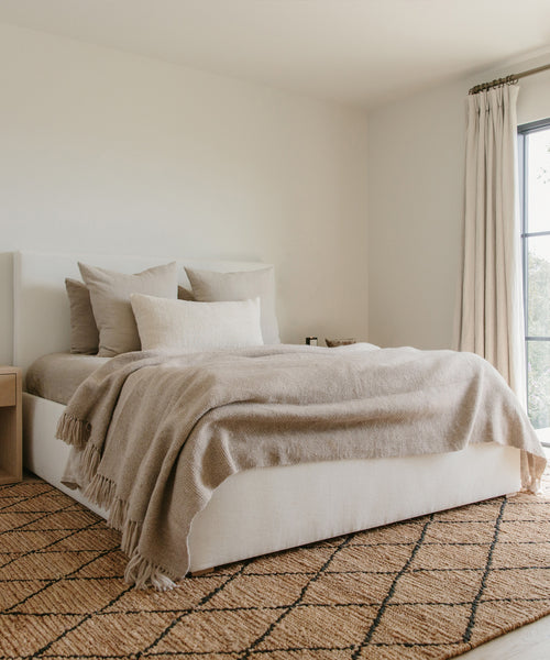 A minimalist bedroom with a neatly made bed, beige bedding and pillows, wooden nightstand, cream curtains, and a patterned brown rug. A Large Alpaca Basketweave Throw adds cozy texture as natural light streams in through the large window.