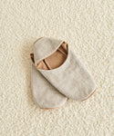 A pair of Linen House Slipper in beige with brown lining, one slightly overlapping the other, placed on a textured cream-colored carpet.