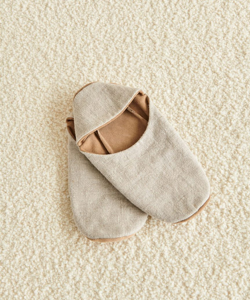 A pair of Linen House Slipper in beige with brown lining, one slightly overlapping the other, placed on a textured cream-colored carpet.