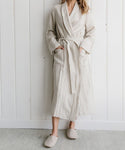 A person stands indoors wearing a long, light beige robe with a belt and matching Linen House Slipper. Hands rest in the robe pockets as they pose before a white, paneled wall, with their head not fully visible.
