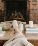 Relaxing with feet up on a wooden coffee table, wearing cozy Linen House Slippers and wrapped in a blanket. The table displays books, a candle, and a bowl, set near a fireplace with stacked logs and a brick wall.