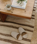 A pair of Linen House Slippers in beige rests on a striped rug beside a wooden coffee table topped with a mug of tea, a small flower vase, and stacked books.