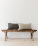 A rustic wooden bench sits against a white wall, topped with two textured rectangular pillows—one dark gray handwoven pillow and one Luna Lumbar Pillow in light beige—on a light wood floor.