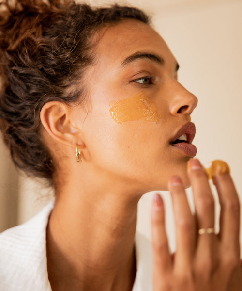 A person in a white robe applies a thick, golden-brown facial mask to their cheek with their fingers. This close-up side profile showcases the Mini Routine—the ideal travel companion for your skincare regimen.