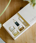 The Mini Routine by Oak Essentials—a cream-colored skincare gift box with five travel-sized products—rests open on a wooden surface, making it the perfect travel companion, with blurred green plants in the foreground.
