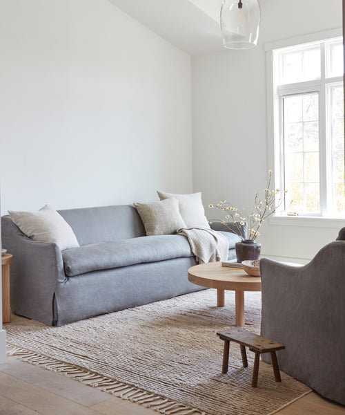 A bright, minimalist living room featuring a gray Miramar Sofa Slipcover, light pillows, a round wood coffee table, textured rug, small wooden bench, and a potted plant against white walls and large windows.