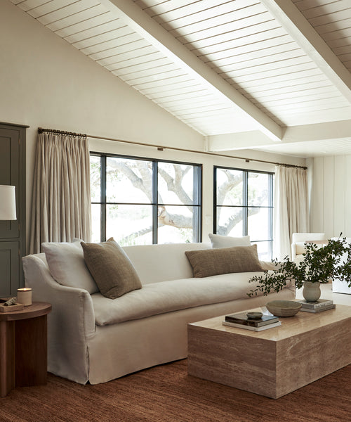 A bright living room features the Miramar Sofa Slipcover in white linen, neutral pillows, a light wood coffee table with a plant and books, large windows with curtains, and exposed white ceiling beams.