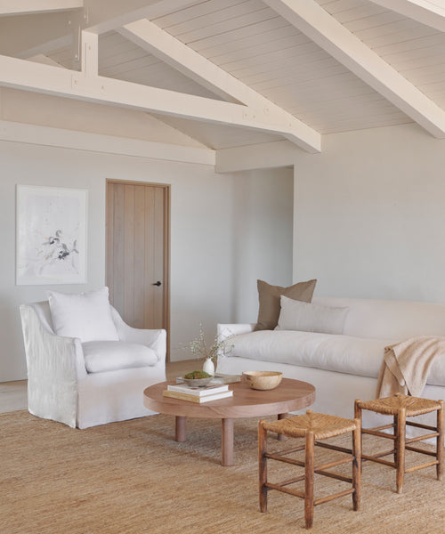 A minimalist living room in neutral tones features the Miramar Chair in white linen, a matching sofa with beige pillows, a round wooden coffee table, woven stools, and a natural fiber rug. Exposed beams and soft lighting add coziness.