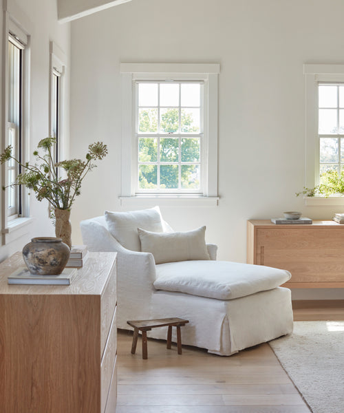 Bright, airy room with large windows, the Miramar Chaise with pillows, light wood furniture, a small wooden stool, a vase of flowers, and pale wood flooring create a minimalist and cozy atmosphere.