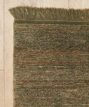 A close-up of the Mojave Jute Rug, featuring earth-toned greenish-brown woven fibers and fringe along the top edge, placed on a light wooden floor.