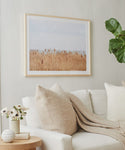 A cozy living room features a white sofa, beige pillows, a knitted throw, a flower-topped side table, and Motion, a Daniella Dolenc photo of golden reeds in a European pine frame, displayed on a light-colored wall.