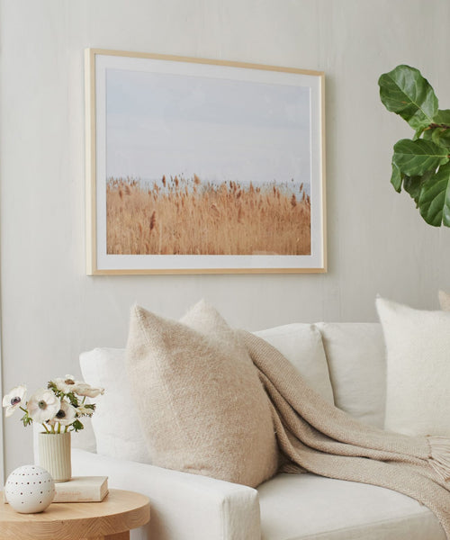 A cozy living room features a white sofa, beige pillows, a knitted throw, a flower-topped side table, and Motion, a Daniella Dolenc photo of golden reeds in a European pine frame, displayed on a light-colored wall.
