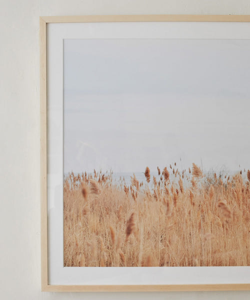 ‘Motion,’ a Daniella Dolenc photograph featuring tall golden-brown grass beneath a pale, cloudy sky, hangs in an elegant European pine frame on a light wall, bringing a peaceful and minimalist touch to the space.