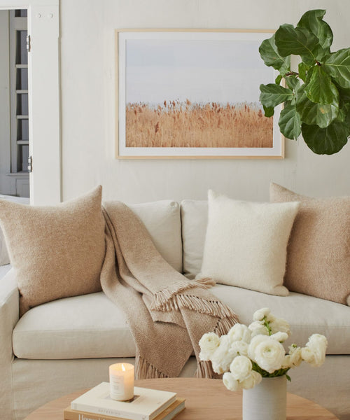 A cozy living room features the Motion light beige sofa, soft pillows, and a tan throw. A candle and white flowers rest on a wooden coffee table, while a framed Daniella Dolenc photograph and leafy plant accent the wall and corner.