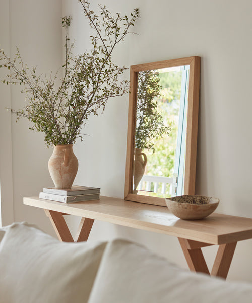 A wooden console table against a white wall displays a vase with leafy branches, books, and a rustic bowl. An Oak Wall Mirror above the table reflects window light. A white sofa sits in the foreground.