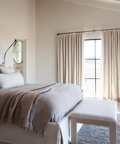 A cozy bedroom with a neatly made bed in neutral bedding, a throw blanket, an Otto Bench at the foot of the bed, beige curtains framing a large window, and soft natural light filling the space.
