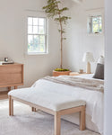 Bright, minimalist bedroom with white walls, a wooden bed and the Otto Bench, light oak furniture, ivory wool boucle bedding, a small table lamp, and a tall potted plant by the window letting in natural light.