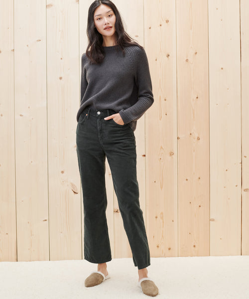 A woman with long dark hair stands relaxed in front of a light wood wall, wearing a dark gray ribbed sweater, Loose Flare Pant in black, and tan slip-on shoes, with one hand casually tucked in her pocket.