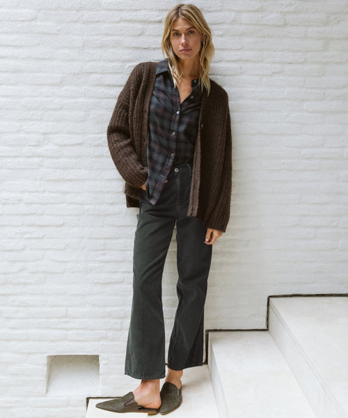 A woman with blonde hair stands casually on white stairs against a white brick wall, wearing Loose Flare Pant, a brown knit cardigan, plaid shirt, and flat shoes.