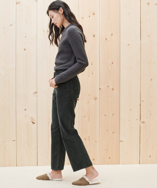 A woman with long dark hair stands in profile against a light wooden wall, wearing a gray ribbed sweater, Loose Flare Pant, and brown slippers with white fleece trim.