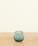 A Roli Poli Glass, small, round, and translucent in smoke blue, is placed on a light wooden surface against a plain off-white background.