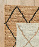 A close-up of two woven rugs overlapping on a wooden floor: the larger is the Wilder Diamond Jute Rug in tan with black diamond patterns; the smaller features a cream-colored diamond design with subtle brown accents.