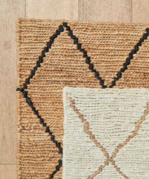 A close-up of two woven rugs overlapping on a wooden floor: the larger is the Wilder Diamond Jute Rug in tan with black diamond patterns; the smaller features a cream-colored diamond design with subtle brown accents.