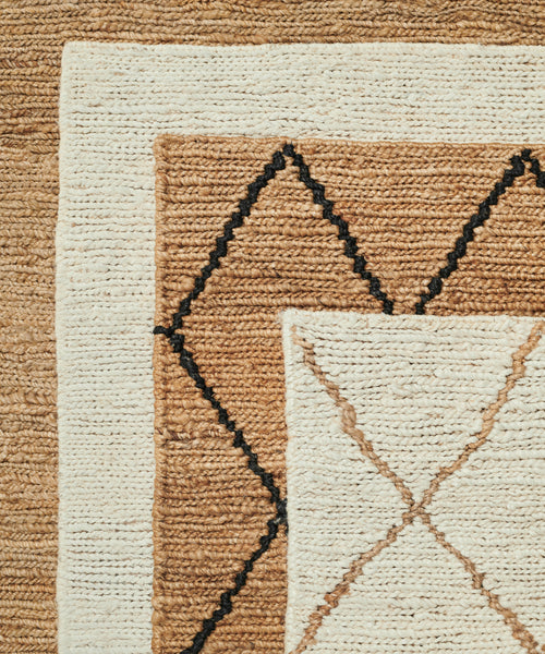A close-up of three overlapping handwoven rugs in neutral tones highlights the Wilder Diamond Jute Rug, which features a bold black and brown diamond pattern on cream and tan with visibly rough, fibrous texture.