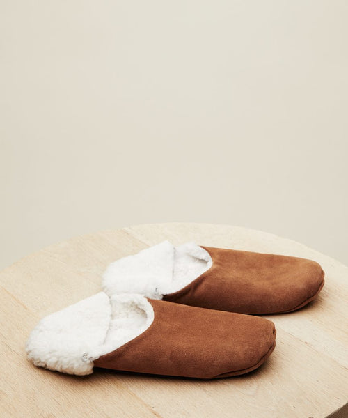 A pair of Suede Moroccan Slippers with shearling lining is displayed side by side on a light wooden surface, set against a plain beige background.