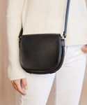 A person in a cream knit sweater and white pants carries the Small Leather Saddle Bag, handmade in Italy from black leather with a curved flap and rope detailing along the edge.