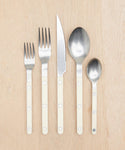 Five utensils—two forks, a knife, a spoon, and a teaspoon—from the Bistro Vintage Finish Cutlery Set with cream handles and silver heads, arranged neatly on a light wooden surface.