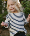 A young child with long blonde hair smiles and looks to the side, wearing the Kids Rue Long Sleeve Tee while standing outdoors with greenery in the background.