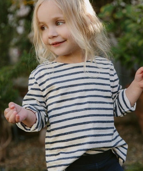 A young child with long blonde hair smiles and looks to the side, wearing the Kids Rue Long Sleeve Tee while standing outdoors with greenery in the background.