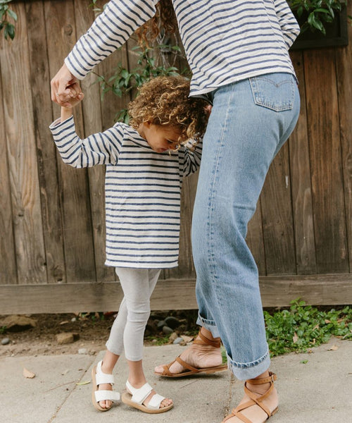An adult and child in matching striped tees, including the Kids Rue Long Sleeve Tee, dance outside by a wooden fence. The child smiles brightly, holding the adults hands as their stripes from Saint James x Jenni Kayne coordinate perfectly.