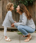 A woman and a young girl smile together outdoors by a wooden fence, both wearing matching Saint James x Jenni Kayne Kids Rue Long Sleeve Tees. The woman crouches, holding the girls hands as they laugh in their striped tees.