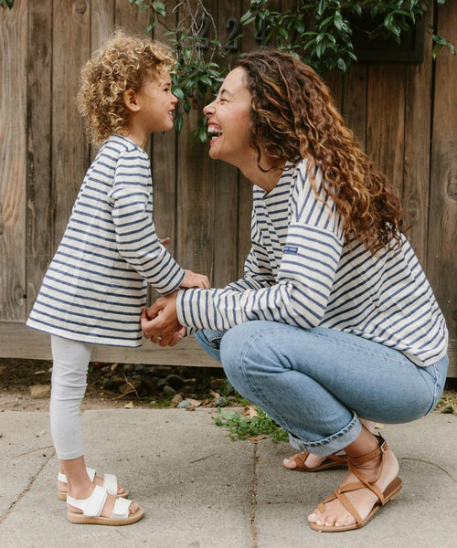 A woman and a young girl smile together outdoors by a wooden fence, both wearing matching Saint James x Jenni Kayne Kids Rue Long Sleeve Tees. The woman crouches, holding the girls hands as they laugh in their striped tees.