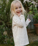 A young girl with long blonde hair, wearing the Kids Rue Long Sleeve Tee, stands outdoors surrounded by greenery and flowers, smiling as she touches her face with one finger.