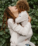 A smiling woman lifts and hugs a happy child in matching light outfits. The child is wearing the Kids Rue Long Sleeve Tee, standing out against a backdrop of lush green leafy plants.