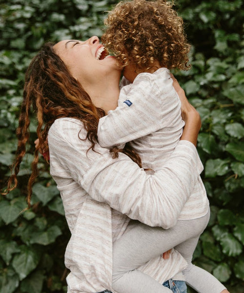 A smiling woman lifts and hugs a happy child in matching light outfits. The child is wearing the Kids Rue Long Sleeve Tee, standing out against a backdrop of lush green leafy plants.