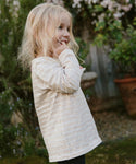 A young blonde girl smiles outdoors, looking to the side, wearing the Kids Rue Long Sleeve Tee, with greenery and potted plants in the background.