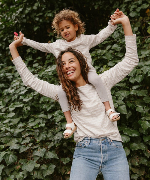 A woman with long curly hair carries a smiling child on her shoulders. The child, wearing the Kids Rue Long Sleeve Tee, holds the woman’s hands as they stand before leafy green plants in their light-colored tops.