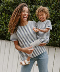 A smiling woman in a Saint James x Jenni Kayne striped shirt and jeans holds a laughing child in the Kids Rue Striped Tee and leggings, outdoors by green bushes and a white fence.