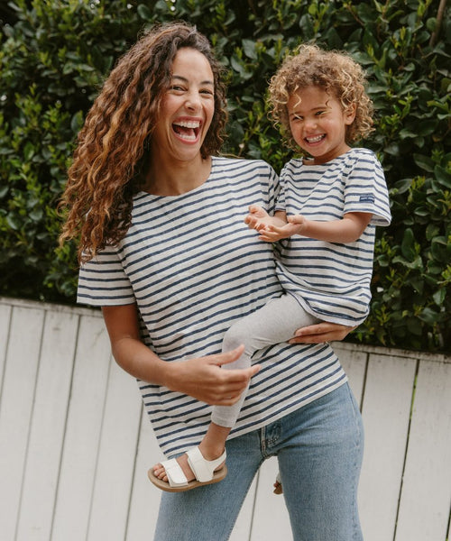 A smiling woman in a Saint James x Jenni Kayne striped shirt and jeans holds a laughing child in the Kids Rue Striped Tee and leggings, outdoors by green bushes and a white fence.