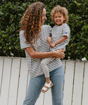 A woman holds a smiling child, both in matching Saint James x Jenni Kayne Rue Striped Tees. The child is wearing the Kids Rue Striped Tee. They stand before a white fence with green bushes behind them.