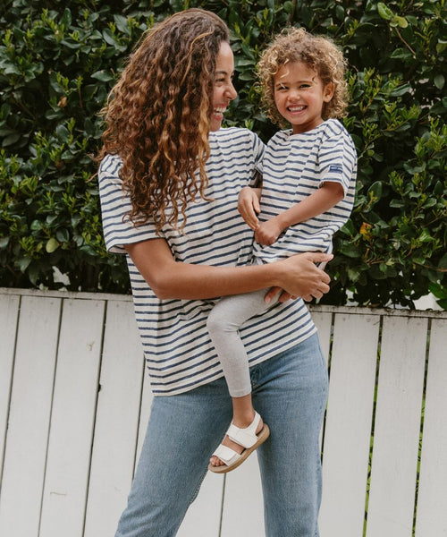 A woman holds a smiling child, both in matching Saint James x Jenni Kayne Rue Striped Tees. The child is wearing the Kids Rue Striped Tee. They stand before a white fence with green bushes behind them.