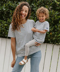 A smiling woman and child stand by a white fence in lush greenery, both wearing Saint James x Jenni Kayne striped shirts. The child is dressed in the Kids Rue Striped Tee with gray leggings and sandals; the woman pairs hers with jeans.