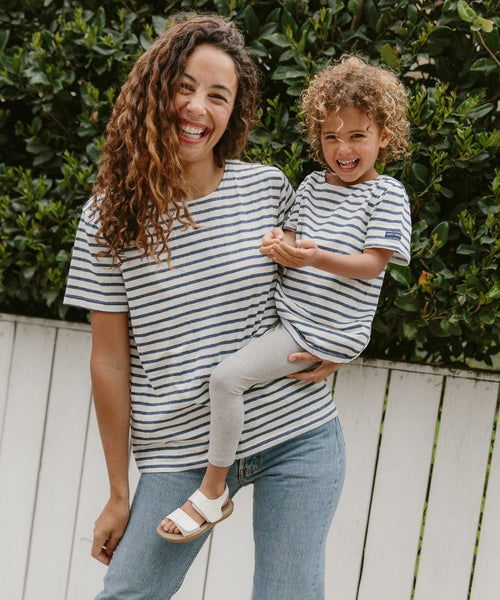 A smiling woman and child stand by a white fence in lush greenery, both wearing Saint James x Jenni Kayne striped shirts. The child is dressed in the Kids Rue Striped Tee with gray leggings and sandals; the woman pairs hers with jeans.